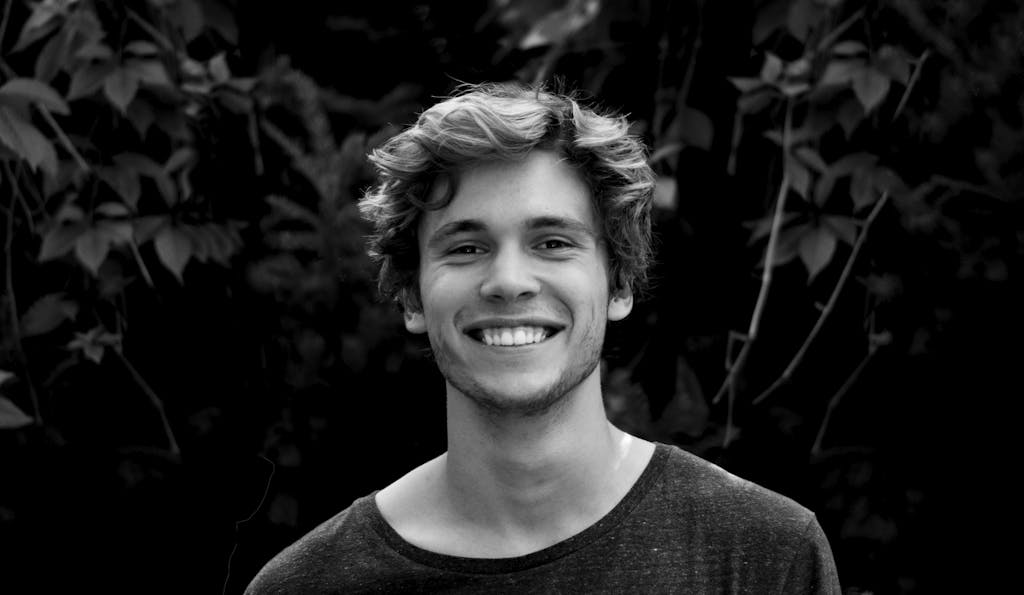 A black and white portrait of a young man smiling outdoors with natural background. He had laser hair removal.