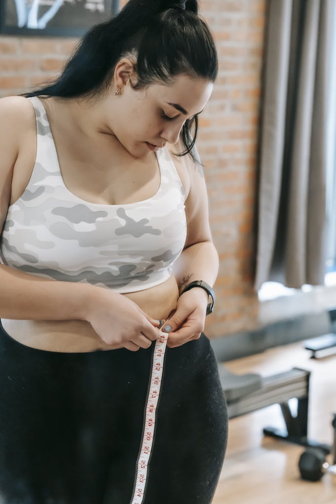 Young obese female using tape for evaluating size of belly while preparing for training in modern fitness center